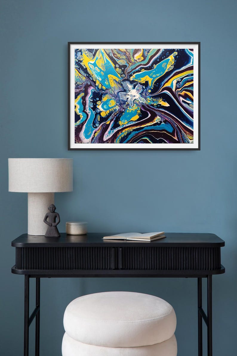 K&ouml;nnte beinhalten: Abstrakter Kunstdruck in einem schwarzen Rahmen. Das Kunstwerk zeigt ein lebendiges, flie&szlig;endes Design mit Wirbeln aus Gelb, Blau, Wei&szlig; und Lila. Der Druck h&auml;ngt &uuml;ber einem schwarzen Schreibtisch mit Lampe, Statue und Buch.