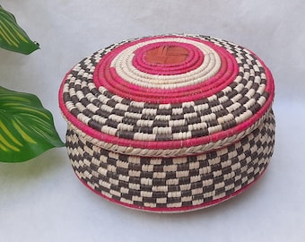 May include: A round, lidded basket with a woven design. The lid features concentric circles of pink, white, and brown, with a central tan circle. The basket's body has a checkerboard pattern of brown and white.