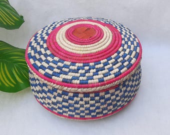 May include: A round, woven basket with a lid. The basket features a checkerboard pattern of blue and white, with pink and red accents. The lid has concentric circles of pink, white, and red. The basket is made of natural fibers.