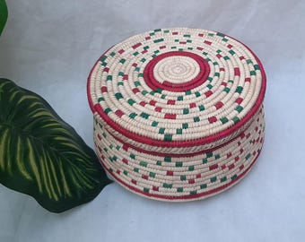 May include: A round, woven basket with a lid. The basket is beige with red and green accents. The lid and the rim of the basket are trimmed in red. The basket is likely used for storage or decoration.