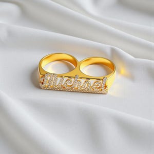 Può includere: Un anello dorato a due dita con il nome Michael in corsivo. La superficie della targhetta è impreziosita da piccole pietre chiare e scintillanti che catturano la luce, su una finitura metallica dorata lucida.
