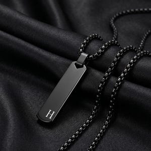 以下が含まれることがあります： 黒色のステンレス鋼ペンダントネックレス。 長方形のペンダントには白い文字「H」があります。 ネックレスチェーンは黒色です。 背景は暗く、質感のある生地です。