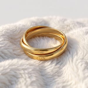 May include: A gold-toned ring composed of three interlocking bands with a smooth, polished finish. The circular design features three distinct, connected loops that overlap to form a single cohesive piece of jewellery with a reflective surface.