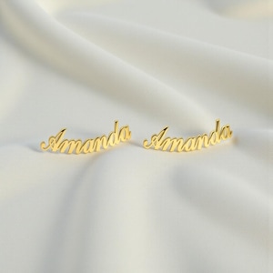 Puede incluir: Dos pendientes dorados, cada uno deletreando el nombre "Amanda" en escritura cursiva. Los pendientes se muestran sobre una superficie blanca texturizada, creando una presentación sencilla y elegante. Los pendientes son una joya personalizada.