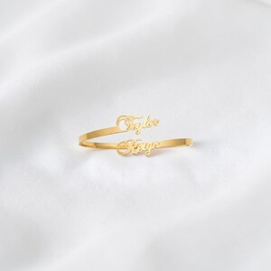 May include: A gold-colored, open-style ring with the names "Taylor" and "Kayla" in a cursive font. The ring is set against a white, textured background, highlighting the jewelry's design and color.