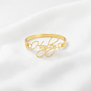 May include: A gold-toned metal bangle bracelet featuring the name Hayley written in a continuous cursive script. The name is integrated into the center of the circular band, which has a smooth, polished finish and a simple, open-ended design.