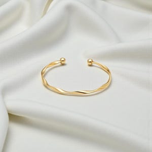 Puede incluir: Un brazalete abierto de color dorado con un diseño retorcido. El brazalete tiene un acabado liso y pulido y pequeños detalles de bola en cada extremo abierto. La joya se exhibe sobre una tela blanca y sedosa.