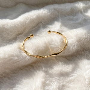 Puede incluir: Una pulsera tipo brazalete de metal dorado con un diseño continuo retorcido y ondulado. La pulsera es abierta y termina con una pequeña cuenta esférica lisa en cada uno de los dos extremos, creando una superficie metálica pulida y brillante.