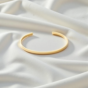 Peut inclure: Un bracelet manchette en métal doré avec une finition lisse et polie, présentant un design simple ouvert aux extrémités. Le bracelet est composé d'une fine bande arrondie qui conserve une largeur constante sur toute sa forme circulaire.