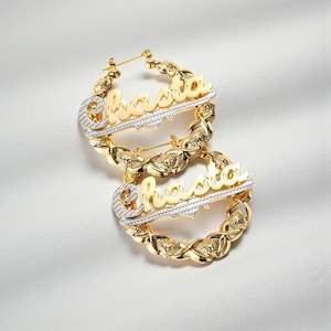 May include: A pair of gold-tone hoop earrings featuring a textured, two-tone design with the name Chasta written in a cursive script across the centre. The hoops have a decorative, repeating pattern along the circular frame with a polished finish.