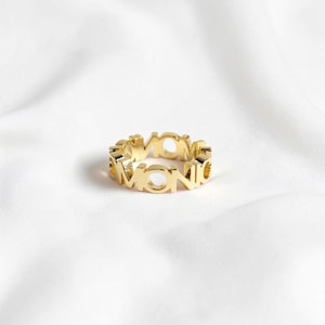 Peut inclure: Une bague dorée présentant un motif découpé répétitif du mot MONIC dans une police de caractères grasse et en bloc. L'anneau possède une surface polie et réfléchissante ainsi qu'une structure ajourée continue formant la forme circulaire du bijou.