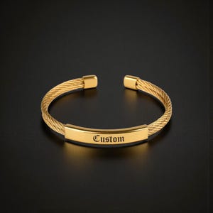 Könnte beinhalten: Ein goldfarbenes, offenes Armband mit einem gedrehten Kabeldrahtband und einer glatten, flachen Mittelplatte. Die Platte ist mit dem Wort Custom in einer schwarzen, gotischen Schrift graviert. Das Armband hat abgerundete Endkappen an der Öffnung.
