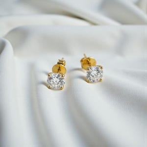 May include: A pair of gold stud earrings, each featuring a round, clear gemstone. The earrings are set in a classic four-prong setting. The background is a white, textured fabric, providing a soft contrast to the jewellery.