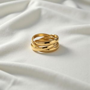 May include: A gold-colored ring with a unique intertwined design. The ring features three bands that are interwoven, creating a stylish and modern look. The ring is set against a white, textured background.