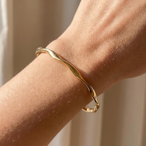 May include: A gold-toned bangle bracelet featuring a smooth, undulating, and wavy surface design. The metal band has a polished, reflective finish that catches the light, creating subtle highlights along its curved, irregular shape.