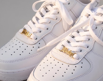 May include: A pair of white sneakers featuring gold-toned metal lace charms. Each charm is shaped like the word Baby in a decorative script font and is threaded onto the white shoelaces at the base of the eyelet panel on each shoe.