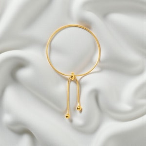 Könnte beinhalten: Ein goldfarbenes, verstellbares Armband mit filigranem Kettendesign. Das Armband verfügt über einen Schiebeverschluss mit Perle und zwei kleine, runde Endperlen. Der Schmuck wird auf einem weißen, seidigen Stoff präsentiert.