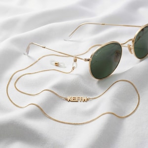 Puede incluir: Gafas de sol doradas con lentes verdes y una cadena a juego. La cadena presenta el nombre "KEITH" en letras de bloque. Las gafas y la cadena descansan sobre una superficie blanca y texturizada.