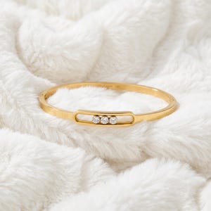 Può includere: Un bracciale rigido in oro caratterizzato da un'apertura ovale orizzontale centrale contenente tre pietre rotonde, chiare e scintillanti disposte in fila. La fascia in metallo ha una finitura liscia e lucida con un profilo sottile.