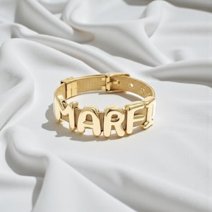 Könnte beinhalten: Ein goldfarbenes Armband mit einem Netzband und großen, abgerundeten, metallisch goldenen Buchstaben, die den Namen MARFI bilden. Das Armband verfügt über einen schnallenartigen Verschluss und ist durchgehend mit einer polierten Oberfläche versehen.