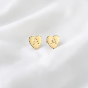 Peut inclure: Une paire de boucles d'oreilles en forme de cœur dorées. Chaque boucle d'oreille porte la lettre "A" en son centre. Les boucles d'oreilles sont présentées sur une surface blanche texturée.