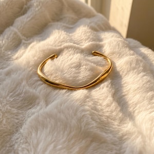 Op de afbeelding: Een goudkleurige metalen armband met een glad, doorlopend en licht onregelmatig golvend ontwerp. De armband heeft een open structuur met afgeronde uiteinden en een gepolijst, reflecterend oppervlak dat het licht opvangt.