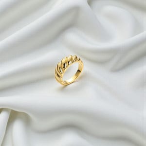 Puede incluir: Un anillo de tono dorado con un diseño continuo, retorcido y similar a un cruasán, con una superficie pulida y reflectante. La banda tiene un perfil grueso y redondeado con profundas ranuras diagonales que crean un patrón texturizado repetitivo.