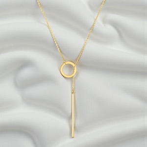 May include: A gold-toned lariat necklace featuring a delicate chain, a circular ring pendant, and a slender vertical bar drop. The necklace is constructed with a polished metal finish, showcasing a clean geometric design with a central ring.