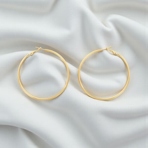 May include: Two gold-colored hoop earrings. The earrings are circular and have a smooth, polished finish. They are displayed on a white, textured fabric background. The earrings are a classic jewelry item.
