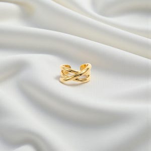 Puede incluir: Un anillo dorado con un diseño entrecruzado. El anillo tiene una banda gruesa y un diseño abierto. El anillo está sobre un fondo blanco y sedoso.