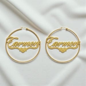 May include: A pair of gold-toned hoop earrings featuring a textured, glittery finish. Each earring displays the name Carmen in a cursive script font, centered within the hoop, with a small heart shape positioned directly below the name.