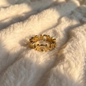 May include: A gold-toned ring featuring a continuous band of cut-out capital letters that spell out the word MONO. The metal surface has a polished, reflective finish, and the design consists of interconnected block-style characters.