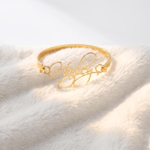 May include: A gold-toned metal bangle bracelet featuring a central script-style nameplate that reads "Hayley". The bracelet has a circular band design with small circular connectors on each side of the nameplate, creating a continuous loop.