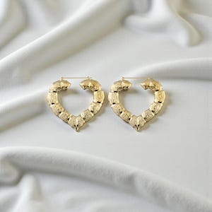 May include: A pair of gold-colored heart-shaped hoop earrings. The earrings have a textured surface and are displayed on a white, draped fabric. The earrings are a fashionable accessory.