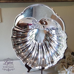 May include: A silver-colored, nautilus-shaped serving dish with a scalloped edge. The dish has a reflective surface and is supported by a dark brown stand. The image also includes the text "Lyon Vintage".