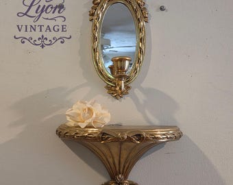 May include: A gold-colored vintage wall shelf with an attached oval mirror and candle holder. The shelf has a decorative bow and leaf design. A cream-colored flower rests on the shelf. The text "Lyon Vintage" is visible.