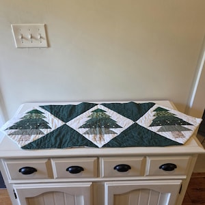 May include: A quilted Christmas tree table runner. The runner features a repeating pattern of green Christmas trees on white diamond-shaped blocks, set against a dark green background. The trees have various patterned fabrics. The runner is placed on a white cabinet.