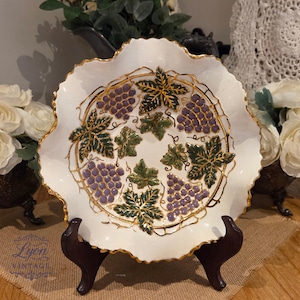 May include: White ceramic decorative plate with a scalloped edge and gold trim. The plate features a design of purple grapes and green leaves with gold accents. The plate is displayed on a dark brown wooden stand.