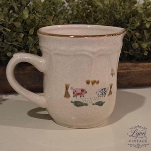 May include: Cream-colored ceramic mug with a brown rim and handle. The mug features a farm scene with two pigs, hay bales, and corn. The mug is a vintage item.