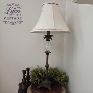 May include: A vintage table lamp with a cream-colored lampshade and a pineapple-shaped glass body. The lamp has a bronze-colored base and stem. The lamp is on a round table with two wooden candle holders and greenery.