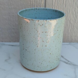 May include: A cylindrical ceramic tumbler featuring a light blue glaze with a speckled finish of small brown dots. The vessel has a smooth, glossy surface texture and a simple, open-top design with straight walls and a flat base.