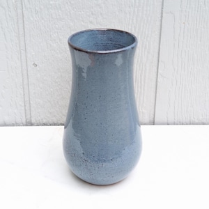 May include: A ceramic vase with a glossy, light blue glaze featuring subtle dark speckles throughout. The vessel has a rounded base that tapers inward toward the middle before widening slightly at the top rim, which has a dark brown edge.