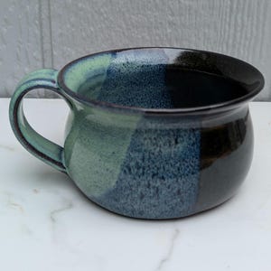 May include: A ceramic mug with a rounded body and a single handle, featuring a glaze that transitions from a light mint green to a speckled blue and finally to a deep, glossy black. The interior and exterior surfaces have a smooth finish.