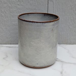 May include: A cylindrical ceramic tumbler featuring a mottled light gray and white glaze with a smooth finish. The vessel has a distinct dark brown rim and a matching brown base, showcasing a simple, functional design with visible clay texture.
