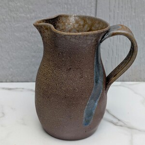 May include: A ceramic pitcher with a textured, dark brown, speckled finish. The vessel features a wide, rounded body, a curved handle, and a prominent pouring spout. A single, vertical streak of blue glaze runs down the side from the handle base.