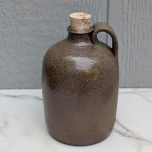 May include: A dark brown stoneware jug with a speckled, textured finish and a single side handle. The jug features a cylindrical body with a narrow neck and is sealed with a light tan, cylindrical cork stopper at the top opening of the vessel.