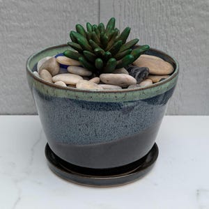 May include: A ceramic planter with a tapered shape and a matching saucer. The pot features a reactive glaze with a dark charcoal base that transitions into a speckled blue and light green rim. A dark green succulent sits atop a bed of smooth river stones.