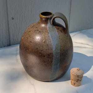 May include: A round, brown ceramic jug with a speckled finish and a single handle. A vertical, light blue-grey glaze stripe runs down the side from the neck. The jug features a narrow opening at the top and is accompanied by a separate cylindrical cork stopper.