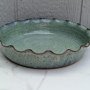 May include: A round ceramic pie dish with a scalloped edge and a speckled light green glaze. The interior and exterior feature a mottled texture with dark brown flecks throughout, and the rim is finished with a dark brown exposed clay edge.