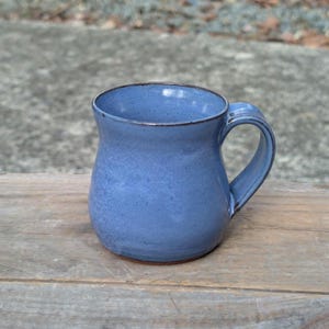 May include: A blue ceramic mug with a rounded body and a wide handle. The mug has a speckled glaze and a dark brown rim.